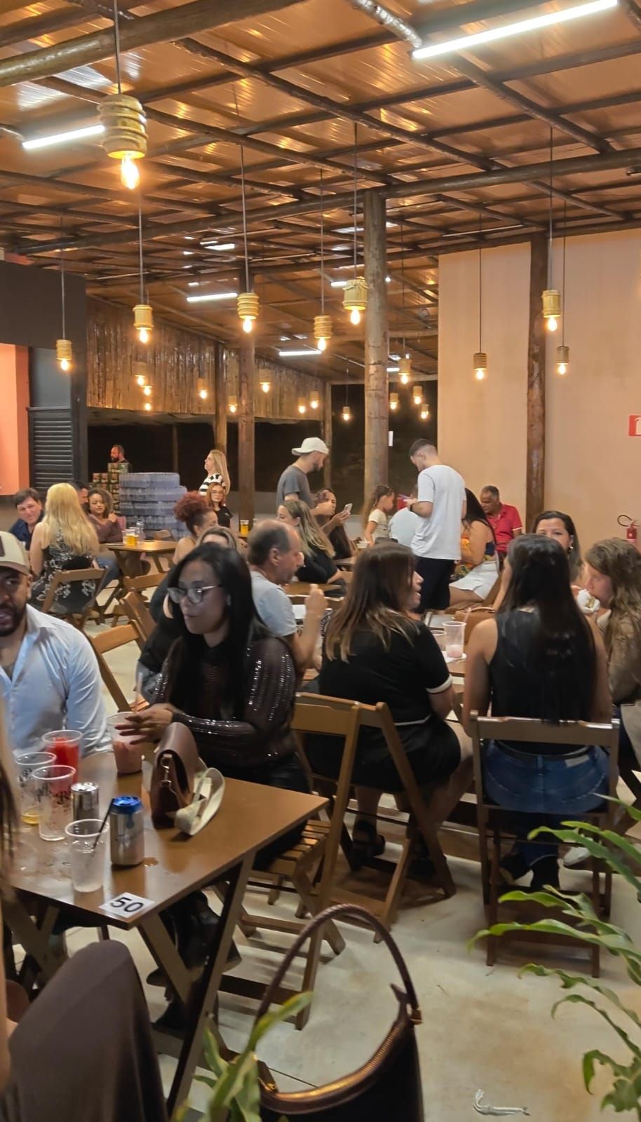 Salão do Vila Bier cheio, com mesas de madeira e luminárias acesas